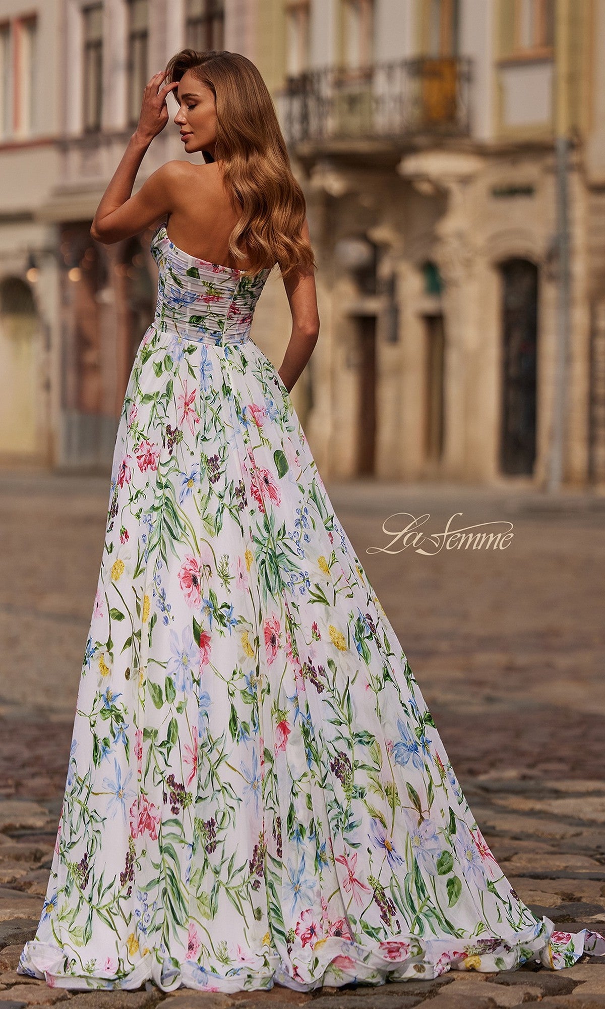 A woman stands outdoors on a cobblestone street, facing away, wearing the La Femme 33450 long prom dress—a strapless floral gown with an a-line skirt and her long wavy hair cascading down her back.